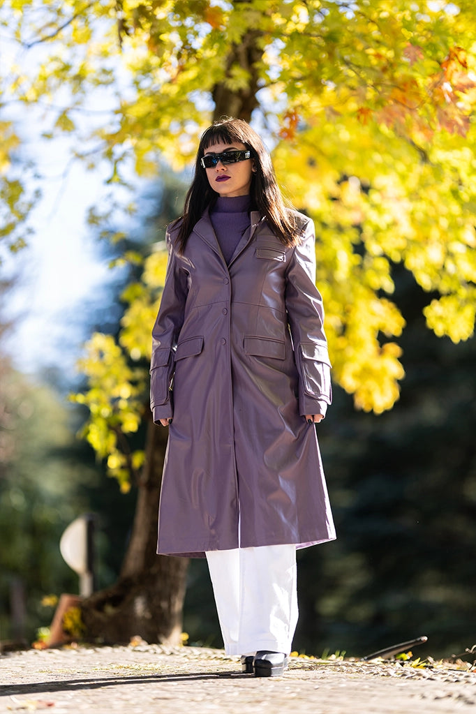 Donna con trench lilla in ecopelle, sfondo alberato giallo autunnale